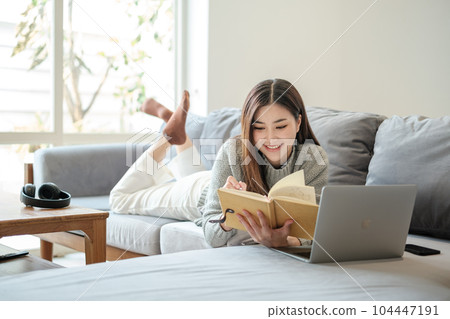 Asian woman woman reading book on sofa in living room. Asian woman woman reading book on sofa in living room. 104447191