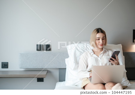Cheerful asian woman looking a message from a Phone Using Laptop Sitting In Bed At Home. Cheerful asian woman looking a message from a Phone Using Laptop Sitting In Bed At Home. 104447195