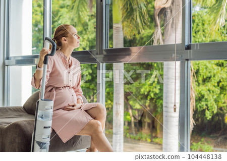 Young pregnant woman enjoys cleaning her house. Easy cleaning with a wireless vacuum cleaner. Pregnant woman cleaning floor with handheld vacuum cleaner. Modern easy cleaning 104448138