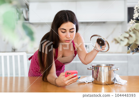 Young woman holding mobile phone opening lid of the pan in the kitchen Young woman holding mobile phone opening lid of the pan in the kitchen 104448393