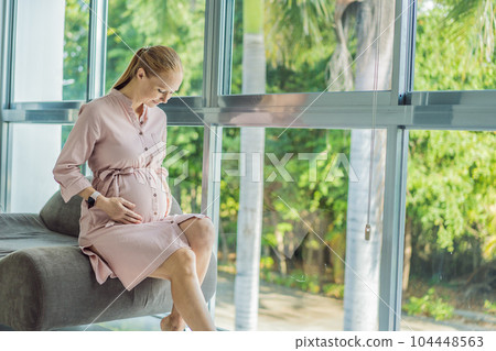 Portrait of young pregnant woman sitting near window holding hands on her belly at modern home with copy space. Pregnancy, motherhood, expectation and tenderness concept 104448563