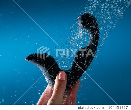 A woman washes a black prostate stimulator under running water on a blue background. Sex toy hygiene concept. 104451837