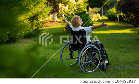 Rear view of an elderly woman spread her arms to the sides while sitting in a wheelchair on a walk outdoors. Rear view of an elderly woman spread her arms to the sides while sitting in a wheelchair on a walk outdoors. 104456817