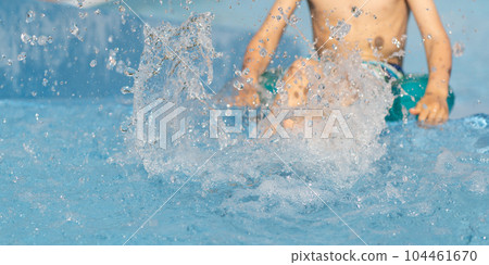 Happy kid splashing water in blue swimming pool on sunny day at summer vacation. Little boy child siting on the edge of the pool, his legs dipping in the water. Copy space 104461670