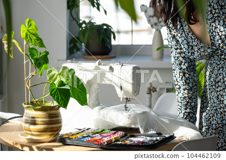A woman sews tulle on an electric sewing machine in a white modern interior of a house with large windows, house plants. Comfort in the house, a housewife's hobby 104462109