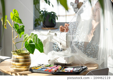 A woman sews tulle on an electric sewing machine in a white modern interior of a house with large windows, house plants. Comfort in the house, a housewife's hobby A woman sews tulle on an electric sewing machine in a white modern interior of a house with large windows, house plants. Comfort in the house, a housewife's hobby 104462119