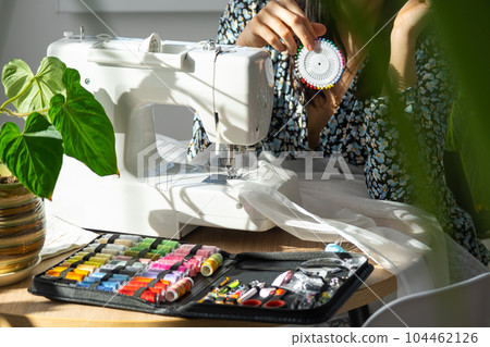 A woman sews tulle on an electric sewing machine in a white modern interior of a house with large windows, house plants. Comfort in the house, a housewife's hobby 104462126