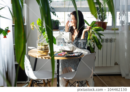 A woman sews tulle on an electric sewing machine in a white modern interior of a house with large windows, house plants. Comfort in the house, a housewife's hobby A woman sews tulle on an electric sewing machine in a white modern interior of a house with large windows, house plants. Comfort in the house, a housewife's hobby 104462130