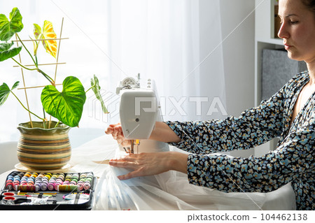 A woman sews tulle on an electric sewing machine in a white modern interior of a house with large windows, house plants. Comfort in the house, a housewife's hobby A woman sews tulle on an electric sewing machine in a white modern interior of a house with large windows, house plants. Comfort in the house, a housewife's hobby 104462138