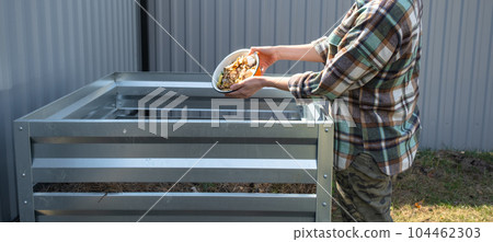 A woman in a plaid shirt pours food waste from a bowl into a compost heap of potato and carrot peelings. Compost box made of metal, eco-friendly fertilizer for the garden A woman in a plaid shirt pours food waste from a bowl into a compost heap of potato and carrot peelings. Compost box made of metal, eco-friendly fertilizer for the garden 104462303