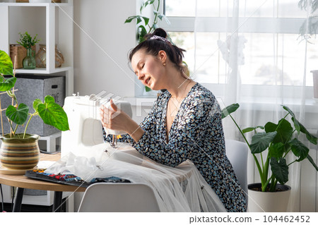 A woman sews tulle on an electric sewing machine in a white modern interior of a house with large windows, house plants. Comfort in the house, a housewife's hobby A woman sews tulle on an electric sewing machine in a white modern interior of a house with large windows, house plants. Comfort in the house, a housewife's hobby 104462452