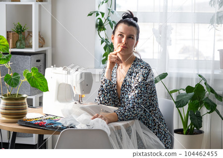Woman thinking, dreaming and sews tulle on an electric sewing machine in a white modern interior of a house with large windows, house plants. Comfort in house, housewife's hobby Woman thinking, dreaming and sews tulle on an electric sewing machine in a white modern interior of a house with large windows, house plants. Comfort in house, housewife's hobby 104462455