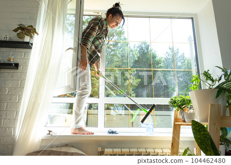 Woman manually washes the window of the house with a rag with spray cleaner and mop inside the interior with white curtains. Restoring order and cleanliness in the spring, cleaning servise 104462480