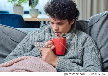 Sick young guy holding blister of pills, lying on couch with mug of hot tea 104465187