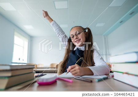 Caucasian girl with two ponytails sits at her desk and raises her hand up to answer the teacher's question. Caucasian girl with two ponytails sits at her desk and raises her hand up to answer the teacher's question. 104465421