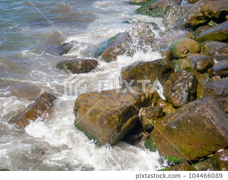 Waves with splashes fall on large stones. Shore view. Beautiful stones that strengthen the shore from the surf. Powerful coastline. Sea foam. The excitement of the sea. 104466089