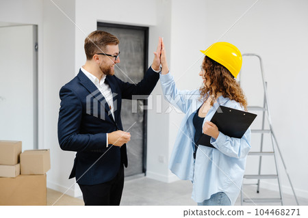 Female designer in helmet with documents giving keys from apartment to young businessman while standing in room Female designer in helmet with documents giving keys from apartment to young businessman while standing in room 104468271