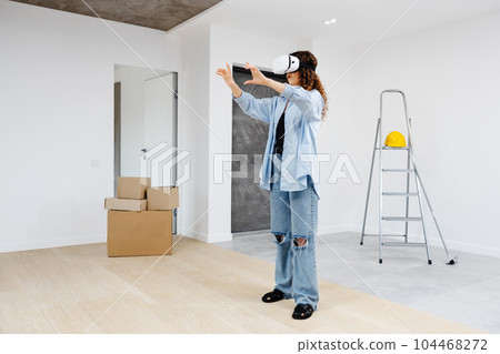 Young woman in VR glasses using modern technology creating room design, moving hands in air, standing in new apartment during renovation. Virtual reality and repair concept 104468272