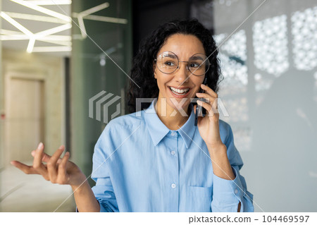 Cheerful and successful mature business woman talking on the phone, hispanic woman smiling and happy sitting inside office at workplace, using laptop at work. 104469597