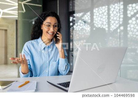 Cheerful and successful mature business woman talking on the phone, hispanic woman smiling and happy sitting inside office at workplace, using laptop at work. 104469598