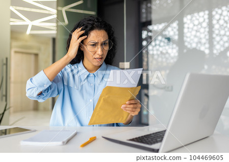 Upset and sad business woman received mail convo letter notification with bad news, financial debt, depressed hispanic woman working inside office building with laptop, reading news. 104469605