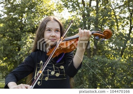 Little Smiling Beautiful Girl Plays On Violin, Musical String Instrument, Green Park And Sun On Background. Little Asian Caucasian Child, Talented Artist, Horizontal Plane. Lifestyle 104470166