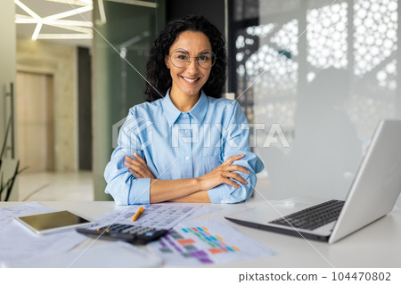 Portrait of successful young businesswoman, Hispanic woman working inside modern office building smiling and looking at camera sitting at desk with laptop, female worker with curly hair and glasses. 104470802