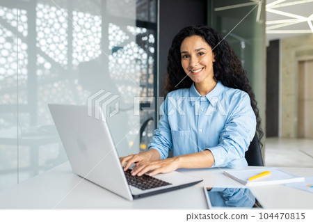 Young beautiful Indian female programmer working inside the office on a laptop, woman smiling and looking at the camera, portrait of a satisfied and confident businesswoman at the workplace. Young beautiful Indian female programmer working inside the office on a laptop, woman smiling and looking at the camera, portrait of a satisfied and confident businesswoman at the workplace. 104470815