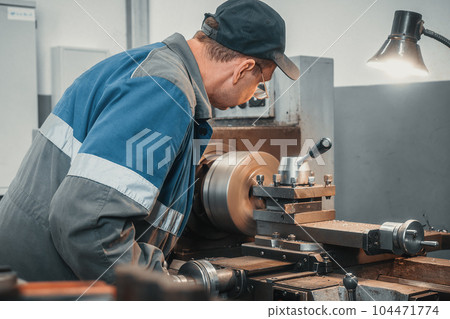 A turner in overalls works on a lathe in a workshop in a factory. Authentic workflow. Back view. A A turner in overalls works on a lathe in a workshop in a factory. Authentic workflow. Back view. A 104471774