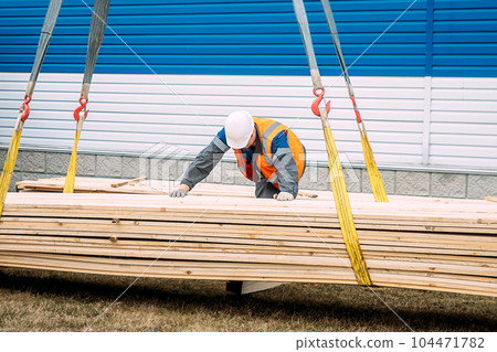 Slinger unloads wooden boards on ground. Worker in helmet and high-visibility vest supervises Slinger unloads wooden boards on ground. Worker in helmet and high-visibility vest supervises 104471782