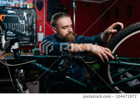 Portrait of bearded cycling mechanic male repairing and fixing mountain bicycle standing on bike rack working in bicycle repair shop with dark interior. Concept of maintenance of bicycle transport. Portrait of bearded cycling mechanic male repairing and fixing mountain bicycle standing on bike rack working in bicycle repair shop with dark interior. Concept of maintenance of bicycle transport. 104471972