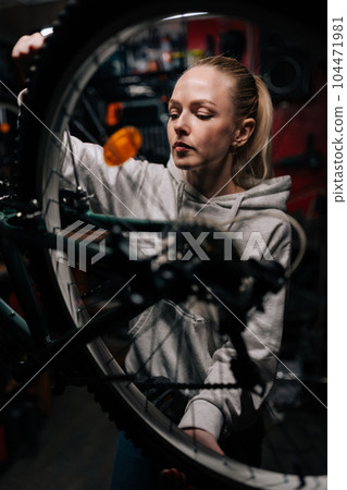Vertical front view of blonde cycling mechanic female repairing and fixing mountain bicycle standing on bike rack in repair workshop with dark interior. Concept of professional bicycle maintenance. 104471981