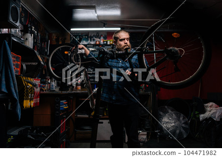 Wide shot portrait of repairman watching rear shifter of mountain bike, changing speeds using handlebar shift lever, working in bicycle repair shop with dark interior. Concept of maintenance of bike. Wide shot portrait of repairman watching rear shifter of mountain bike, changing speeds using handlebar shift lever, working in bicycle repair shop with dark interior. Concept of maintenance of bike. 104471982