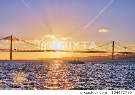 View of 25 de Abril Bridge famous tourist landmark of Lisbon connecting Lisboa and Almada on Setubal Peninsula over Tagus river with tourist yacht silhouette at sunset. Lisbon, Portugal View of 25 de Abril Bridge famous tourist landmark of Lisbon connecting Lisboa and Almada on Setubal Peninsula over Tagus river with tourist yacht silhouette at sunset. Lisbon, Portugal 104472789