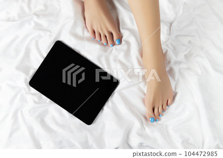 Womans feet with minimal nail design and black tablet on white bed. Beauty salon concept 104472985