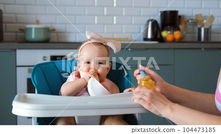 Adorable baby girl with bow on head eating vegetable puree in high chair in the kitchen Adorable baby girl with bow on head eating vegetable puree in high chair in the kitchen 104473185