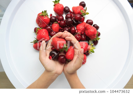 Hands hold large red berries against a white table with strawberries, cherries and pomegranates. 104473632