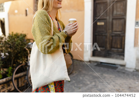 Young Girl holding cup of coffee and white cotton tote bag on old town background. Mockup and zero waste concept. Eco Nature Friendly Style. Environmental Conservation Recycling mock up Young Girl holding cup of coffee and white cotton tote bag on old town background. Mockup and zero waste concept. Eco Nature Friendly Style. Environmental Conservation Recycling mock up 104474452