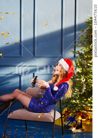 happy woman in shiny blue dress in santa's hat with champagne by Christmas tree 104475610