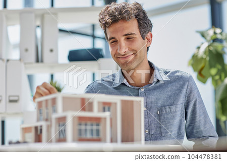 young caucasian engineer man making building model with the use of drawings and notes in office. smart designer assembles a complex structure young caucasian engineer man making building model with the use of drawings and notes in office. smart designer assembles a complex structure 104478381