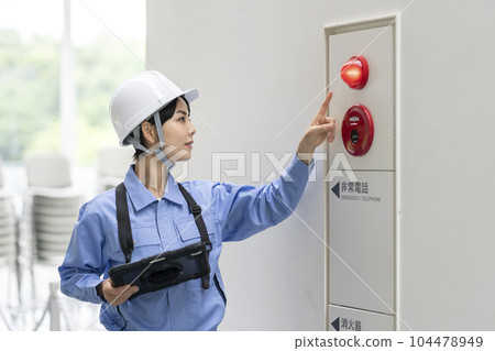 A woman in work clothes inspecting a fire alarm building maintenance image 104478949