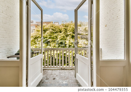 an open door leading to a balcony with white brick walls and green trees in the distance, on a sunny day 104479355