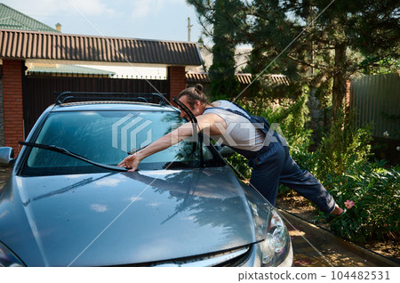 Handsome muscular man washing his car with a high pressure cleaning equipment in the backyard, cleaning wipers. Water compressor. High pressure cleaning equipment. Car detailing. Hand washer, cleaner 104482531
