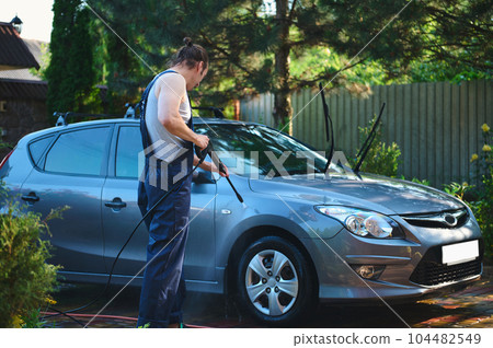 Full-length portrait. Car serviceman in blue work uniform, cleaning a vehicle with a high pressure water jet outdoors. High pressure water cleaner for manual car washing. People Technologies Transport Full-length portrait. Car serviceman in blue work uniform, cleaning a vehicle with a high pressure water jet outdoors. High pressure water cleaner for manual car washing. People Technologies Transport 104482549
