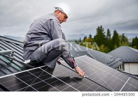 Man technician mounting photovoltaic solar panels on roof of house. Engineer in helmet installing solar module system with help of hex key. Concept of alternative, renewable energy. Man technician mounting photovoltaic solar panels on roof of house. Engineer in helmet installing solar module system with help of hex key. Concept of alternative, renewable energy. 104485300