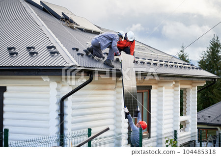 Men mounters installing solar panel system on roof of house. Workers in helmets lifting up photovoltaic solar module outdoors. Concept of alternative and renewable energy. 104485318