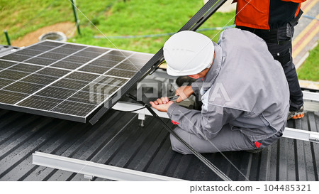Mounters connecting cables while installing photovoltaic solar panels on roof of house. Workers mounting of PV solar modules on rooftop. Concept of alternative and renewable energy. Mounters connecting cables while installing photovoltaic solar panels on roof of house. Workers mounting of PV solar modules on rooftop. Concept of alternative and renewable energy. 104485321