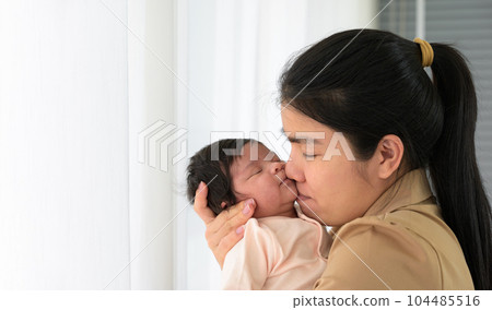 Asian young mom kissing her cute newborn little baby girl sleeping on mom arms near window at home 104485516