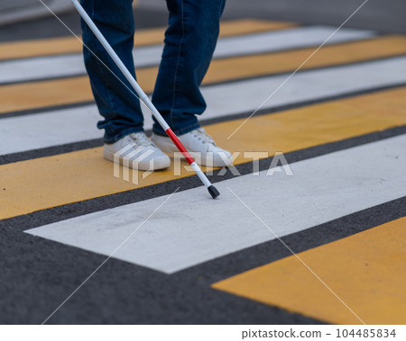 Close-up of the legs of a blind woman crossing the road at a crosswalk with a cane.  104485834
