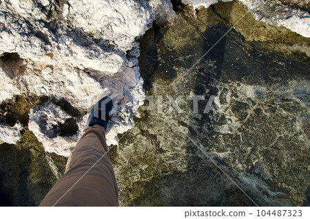 MELLIEHA, MALTA - JANUARY 01, 2020: A man's foot crosses small rock pool with salt water on a rocky coast of Malta near Coral Lagoon MELLIEHA, MALTA - JANUARY 01, 2020: A man's foot crosses small rock pool with salt water on a rocky coast of Malta near Coral Lagoon 104487323